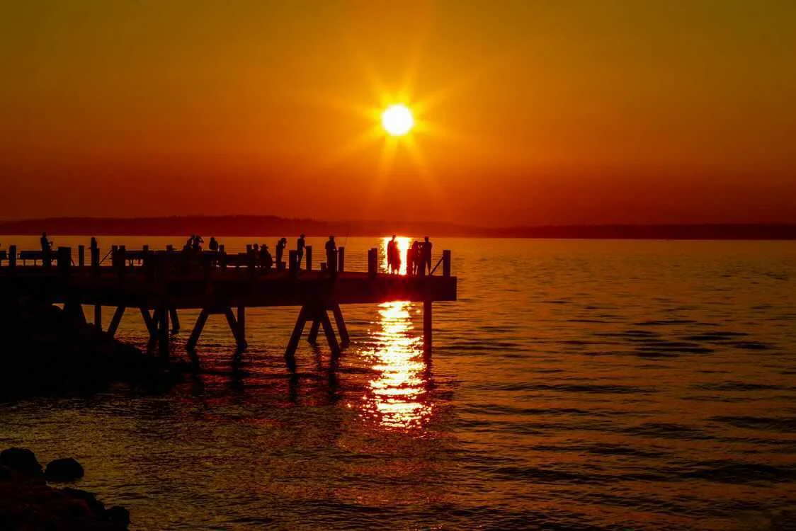 Silhouettes on a wooden pier beneath a blazing orange sunset over calm water — fine art print